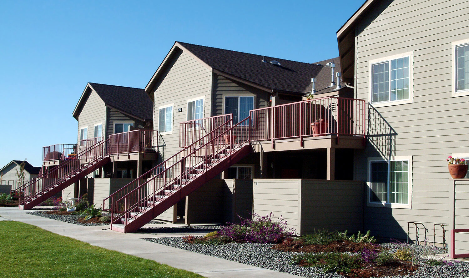 A row of two-story apartment buildings with beige siding and red staircases on a sunny day. Each unit has a small balcony with potted plants. A well-maintained lawn and a sidewalk run parallel to the buildings.