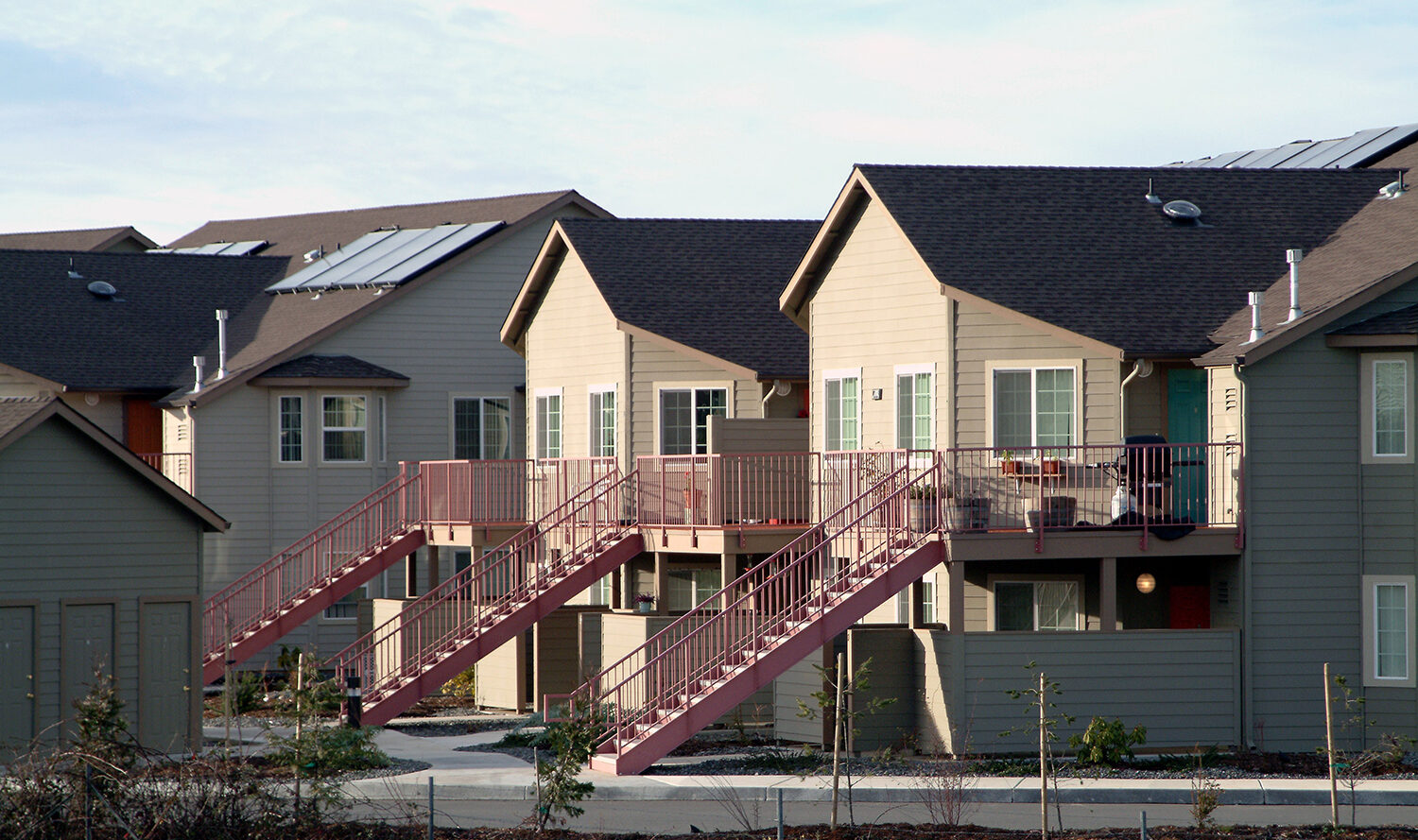 A row of beige, two-story apartment buildings with brown roofs. Each unit has a staircase leading to the upper floor, and the buildings are surrounded by a small landscaped area. Solar panels are visible on some roofs.