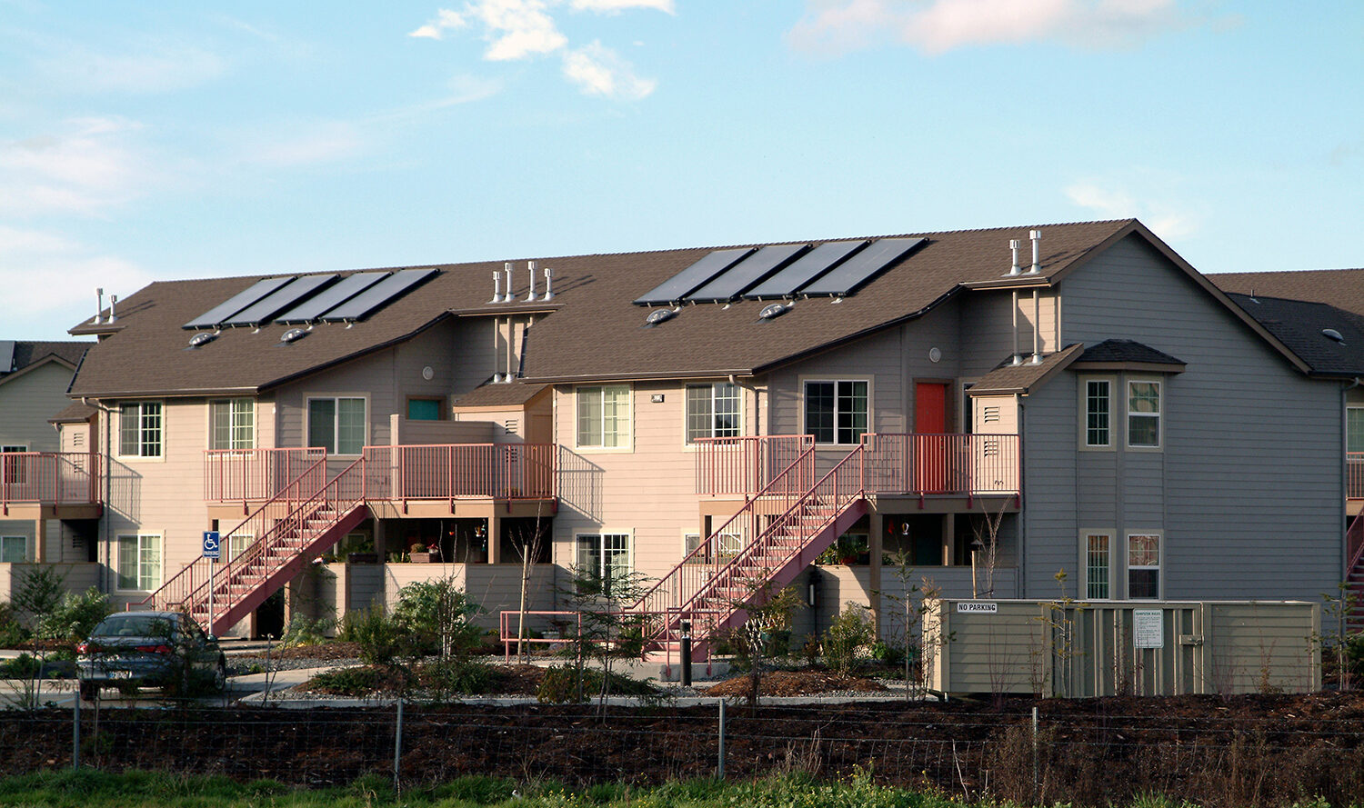 A row of modern townhouses with solar panels on the roof. The buildings are light gray with pink staircases leading to second-floor entrances. A car is parked nearby, and a small grassy area with a fence is in the foreground.