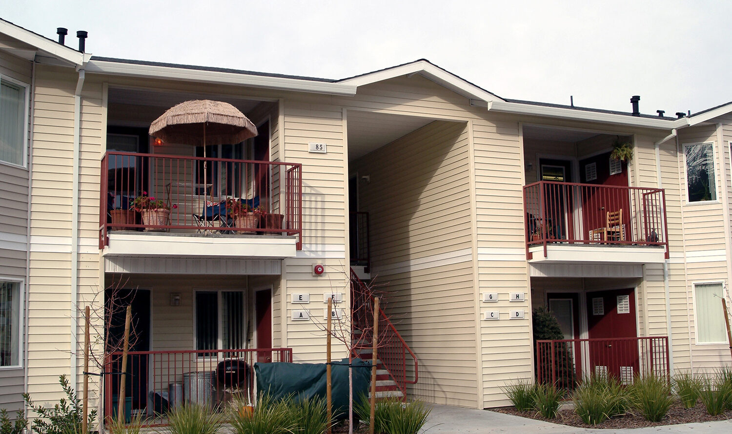A beige two-story apartment building with balconies on the second floor. The left balcony has a large patio umbrella and furniture. A central staircase leads to the upper level. Small plants and shrubs are in front of the building.