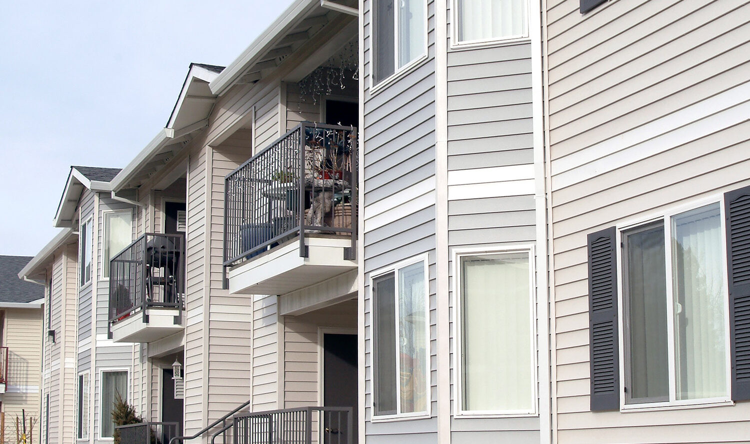Exterior view of a modern apartment building with gray siding and white trim. Several balconies feature small decorations. A curved concrete path winds through a landscaped area with plants and mulch in front of the building.