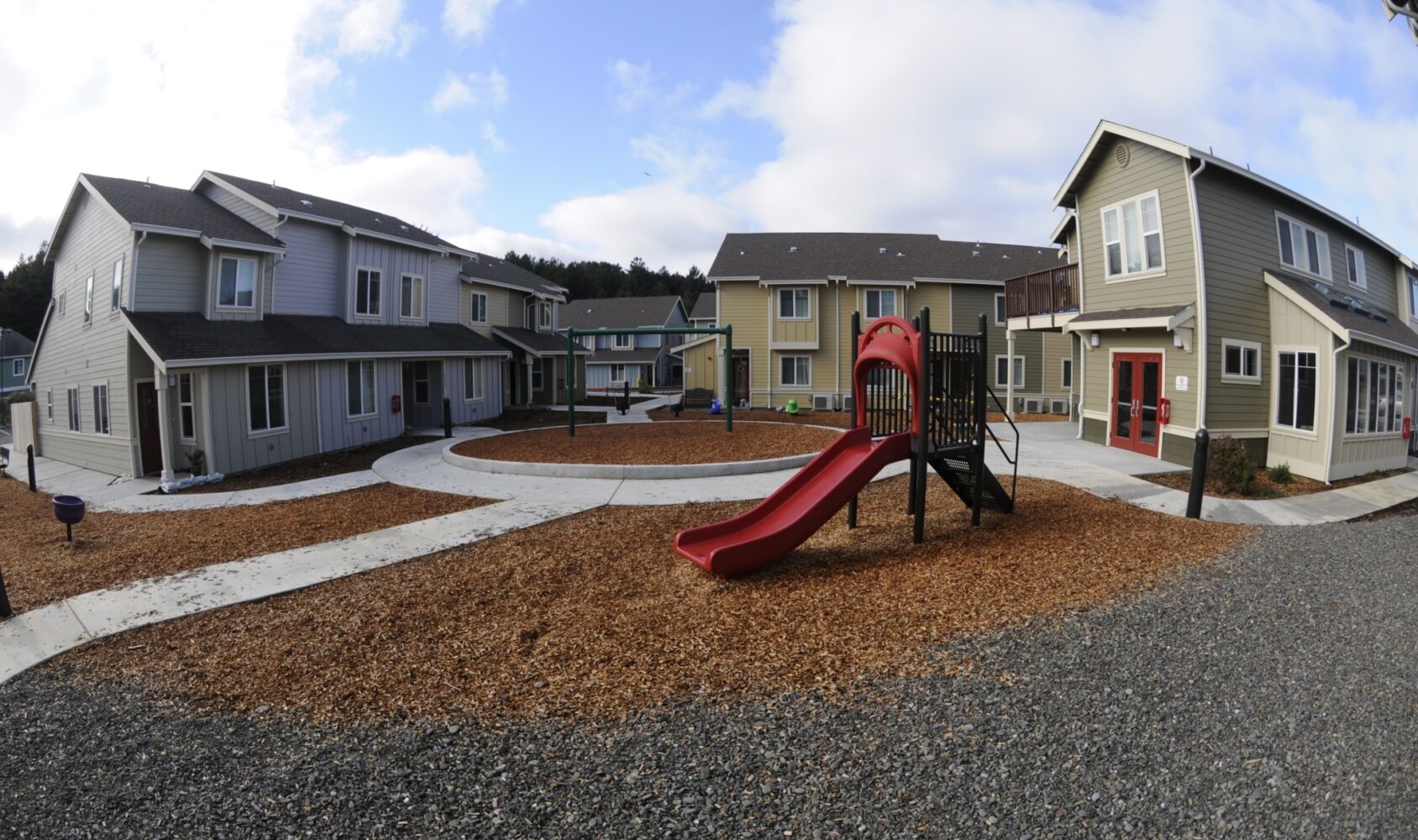 A community area with several two-story houses. A red playground slide is in the center, surrounded by wood chips. Curved sidewalks connect the homes. The sky is partly cloudy, and trees are visible in the background.