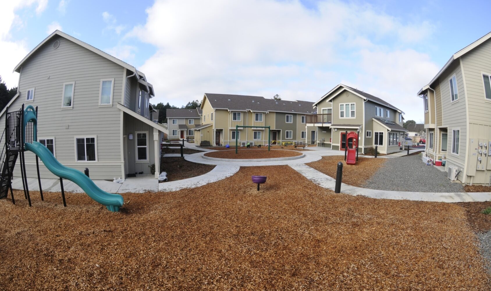A courtyard with woodchip paths is surrounded by three two-story houses. The area features a green slide, a small red playground structure, and open spaces under a partly cloudy sky.
