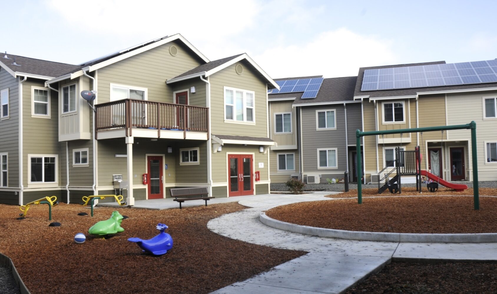 A two-story community housing complex with solar panels on the roof. In front, theres a playground with slides and colorful seesaws, surrounded by a path and mulch. A sunny day with a clear sky.