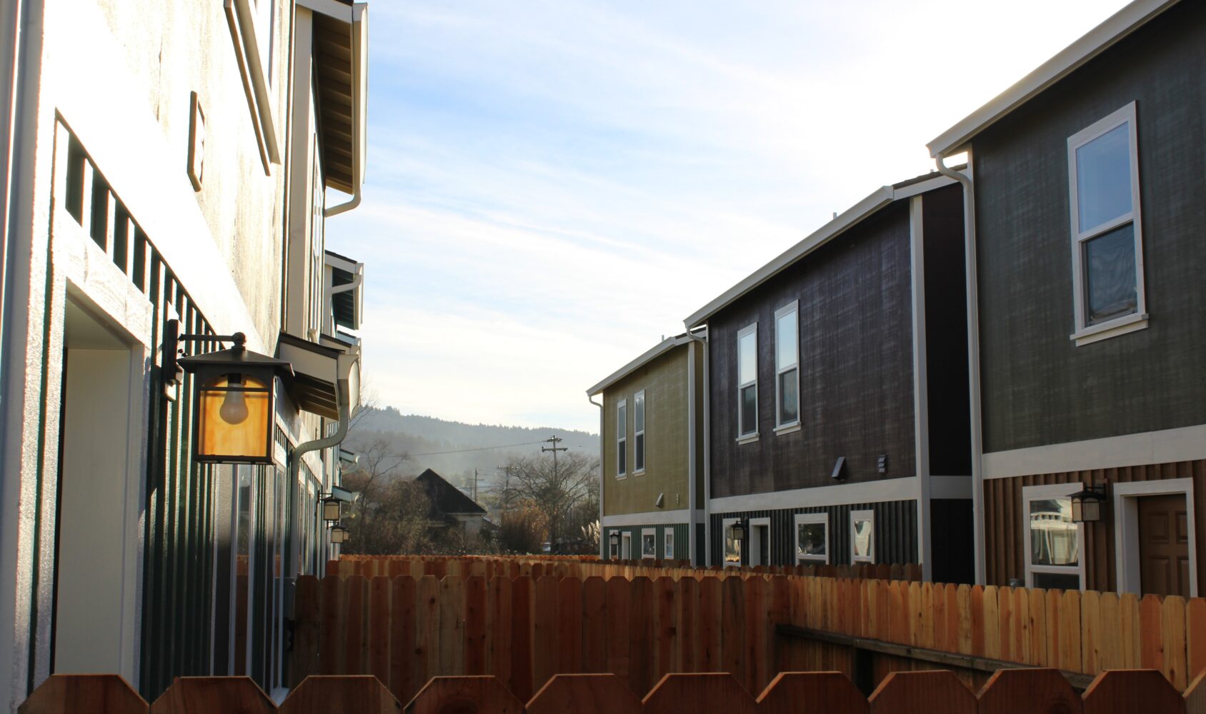 A narrow alley between two rows of suburban houses. A rustic wooden fence lines the path, and warm sunlight illuminates the lamp on one house. The sky is clear, with distant hills in the background.