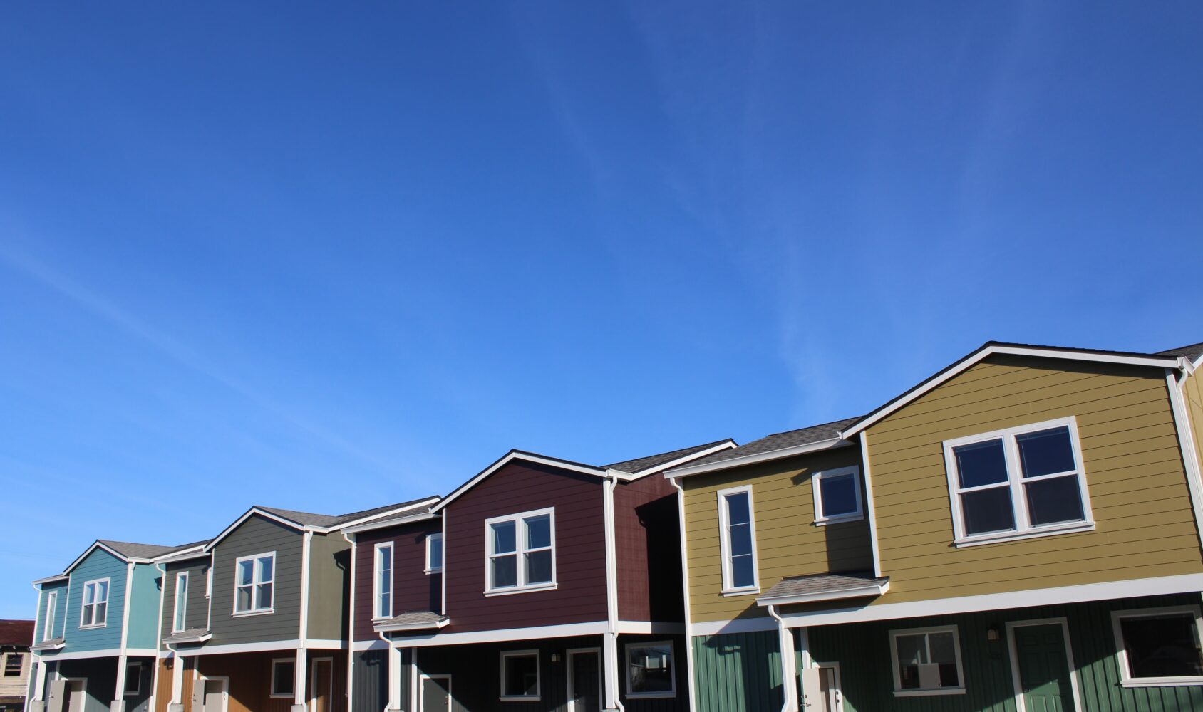 A row of modern, colorful townhouses under a clear blue sky. The homes feature varied facades with different colors including blue, red, and green. The street is empty, creating a peaceful suburban atmosphere.