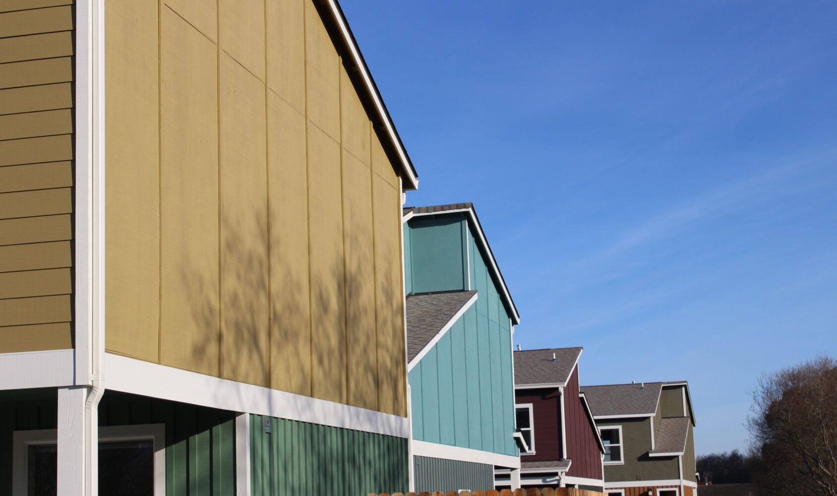 A row of colorful two-story houses with yellow, green, teal, and maroon exteriors are lined up under a clear blue sky. A wooden fence borders the houses, and tree shadows are cast on the yellow house.