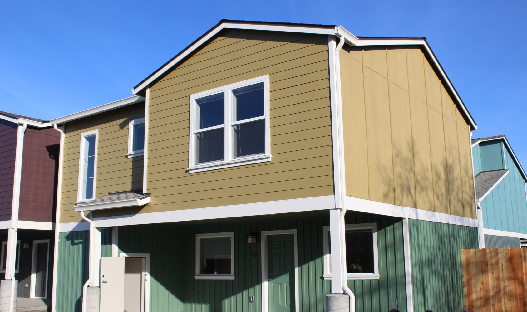 A modern two-story house with a green and mustard-yellow exterior. It has a covered entrance, multiple windows, and is part of a row of similar homes. The sky is clear and blue.