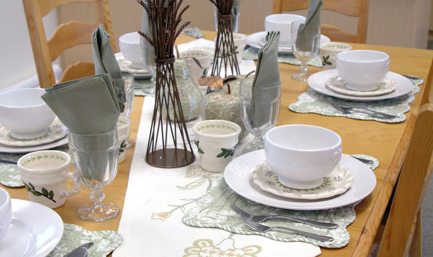 A dining table is set for a meal with white plates, bowls, and green napkins. Tall green candles and decorative centerpieces adorn the table. The table is surrounded by wooden chairs, and a white kitchen is visible in the background.