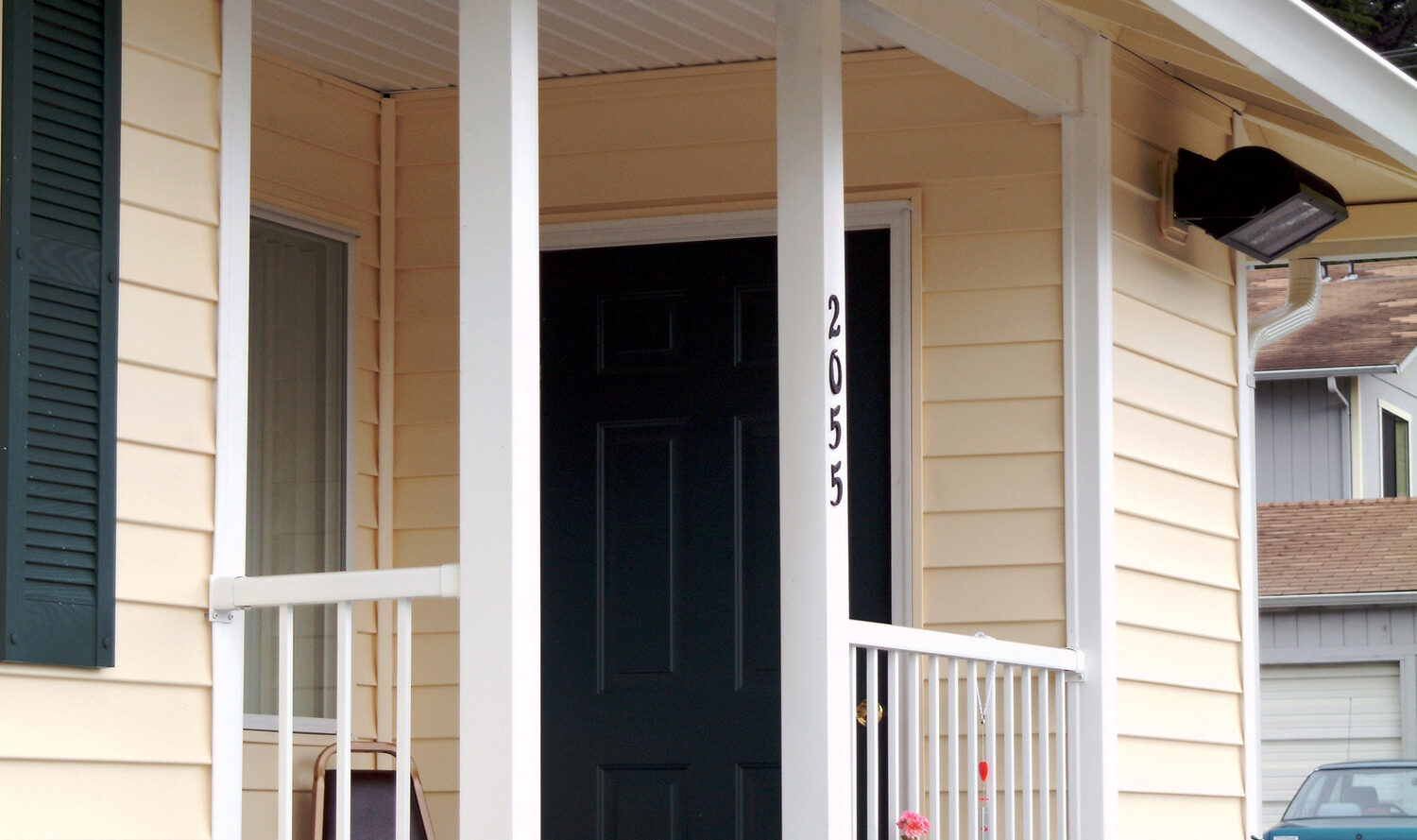 A cozy beige house with a dark front door. The entrance features a small covered porch with white railings. Several potted plants with flowers line the walkway among pebbles, adding a touch of greenery. A parked car is visible in the driveway.