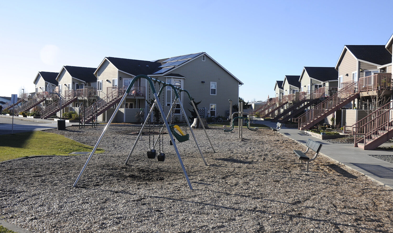 A residential area with multiple two-story townhouse-style homes. A central playground features swings and is surrounded by gravel. Staircases lead to the upper floors of the houses, and a clear sky is visible.