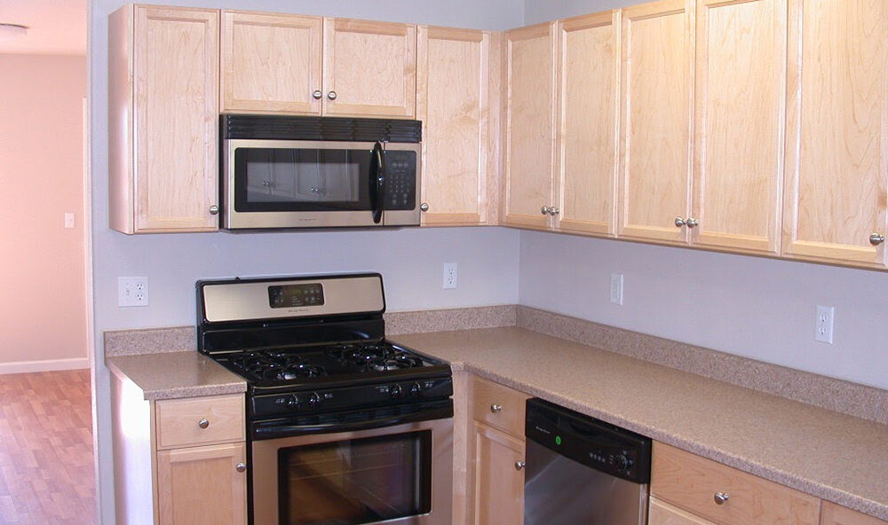 A modern kitchen with light wood cabinets, a black microwave, a black and stainless steel oven, and a stainless steel dishwasher. The countertops are beige, and the flooring appears to be light hardwood.