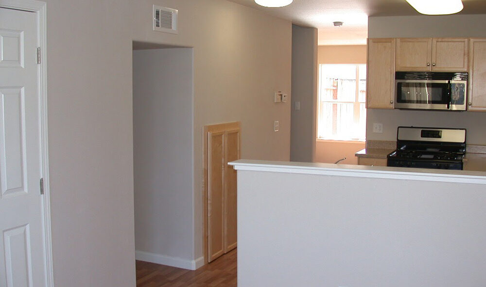 A modern kitchen with light wooden cabinets, a black oven, and microwave. There is a white partition wall with a power outlet. The floor is wooden, and there’s a window allowing natural light. The walls are painted beige.
