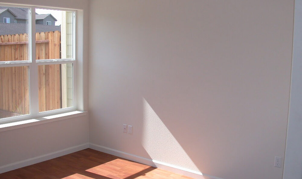 A bright, empty room with light hardwood floors. Sunlight streams in through a large window on the left, casting a shadow on the floor. The walls are painted white, with a fence and neighboring house visible through the window.