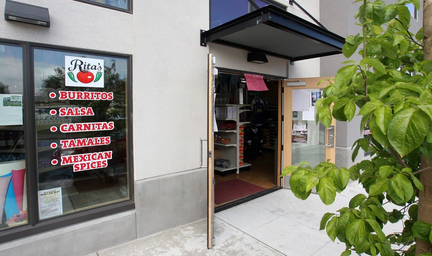 A storefront with a sign for Ritas next to a tree. The window displays menu items: burritos, salsa, carnitas, tamales, and Mexican spices. The entrance door is open, revealing part of the interior.