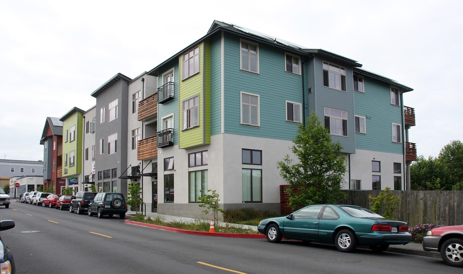 A modern, three-story apartment building with green and gray exterior panels along a street lined with parked cars. The sky is overcast, and a small tree is visible near the building.
