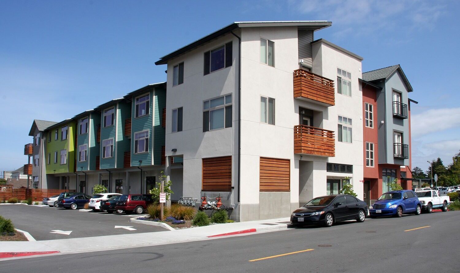 Modern three-story apartment building with colorful facades, featuring balconies and wooden accents. Cars are parked along the street, and the sky is clear. The architecture includes a mix of green, blue, and neutral tones.