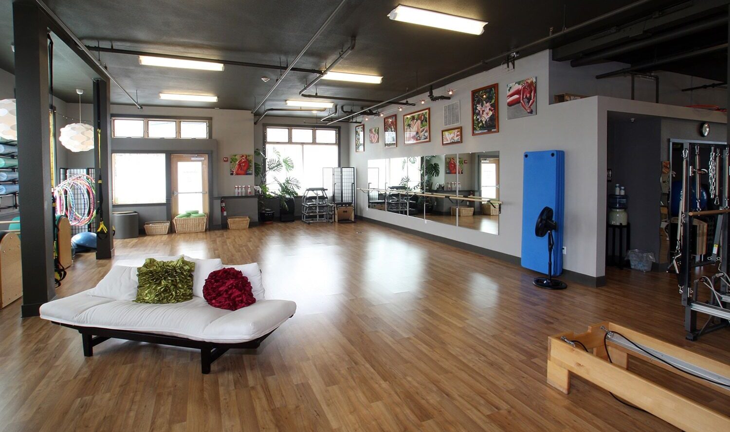 A spacious exercise studio with wooden flooring, a large mirror on the wall, and various exercise equipment. A white futon with red and green pillows is in the center. Wall art and natural light from windows create a vibrant atmosphere.