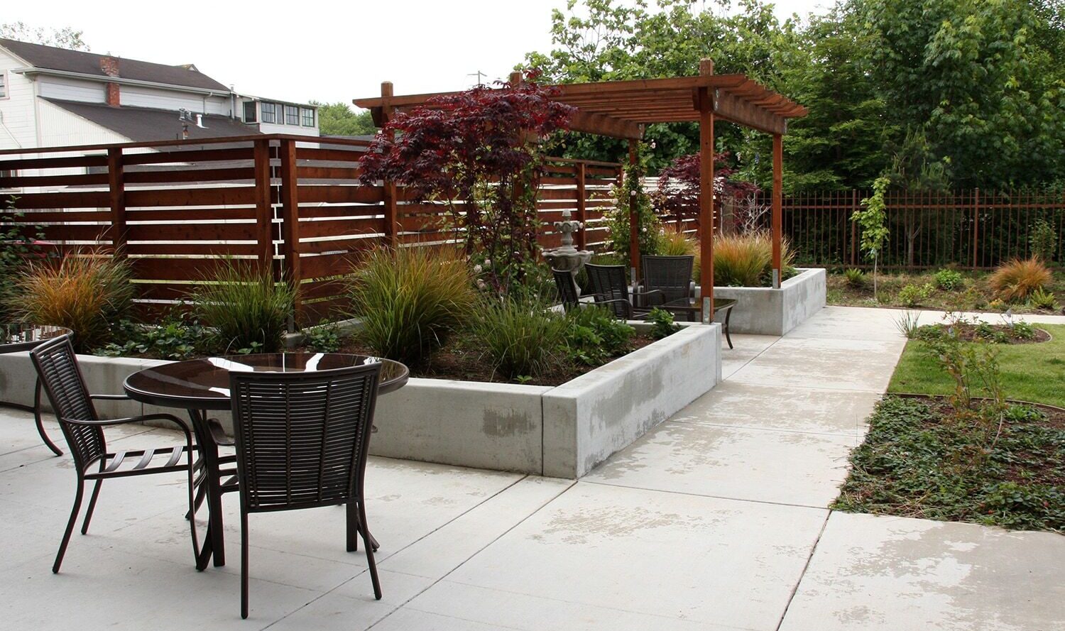 A modern backyard patio with a pergola, concrete planters, and outdoor seating. The space features greenery and ornamental grass. A round table with chairs is in the foreground, and a wooden slat fence encloses the area.