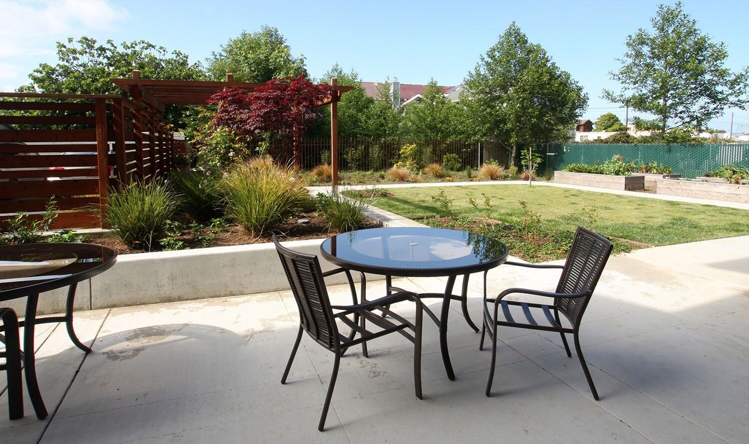 A modern patio with glass-topped tables and metal chairs, overlooking a landscaped garden with green lawn, trees, and shrubs. A wooden pergola stands to the left, offering partial shade. The sky is clear and blue.