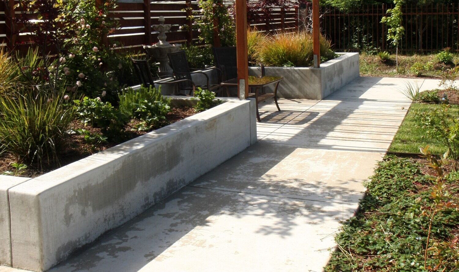 A serene garden pathway with a wooden pergola, surrounded by lush greenery and low plants. The path is made of concrete slabs, with benches under the pergola for relaxing. The background features a wooden fence and trees under a partly cloudy sky.