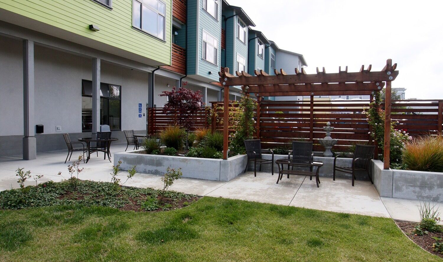 A courtyard featuring a wooden pergola over a paved area with several chairs. There are tables with chairs on the left and landscaped greenery around the concrete paths. Modern building exteriors are visible in the background.