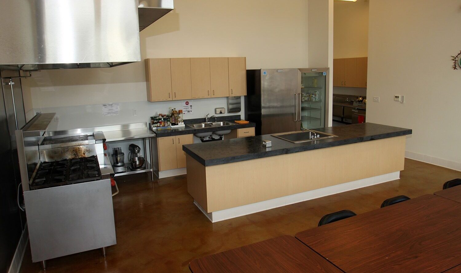 A commercial kitchen featuring a central island with a sink, wooden cabinets, stainless steel appliances, and a large stove. Theres also a long wooden table with chairs in the foreground. The space is clean and well-organized.