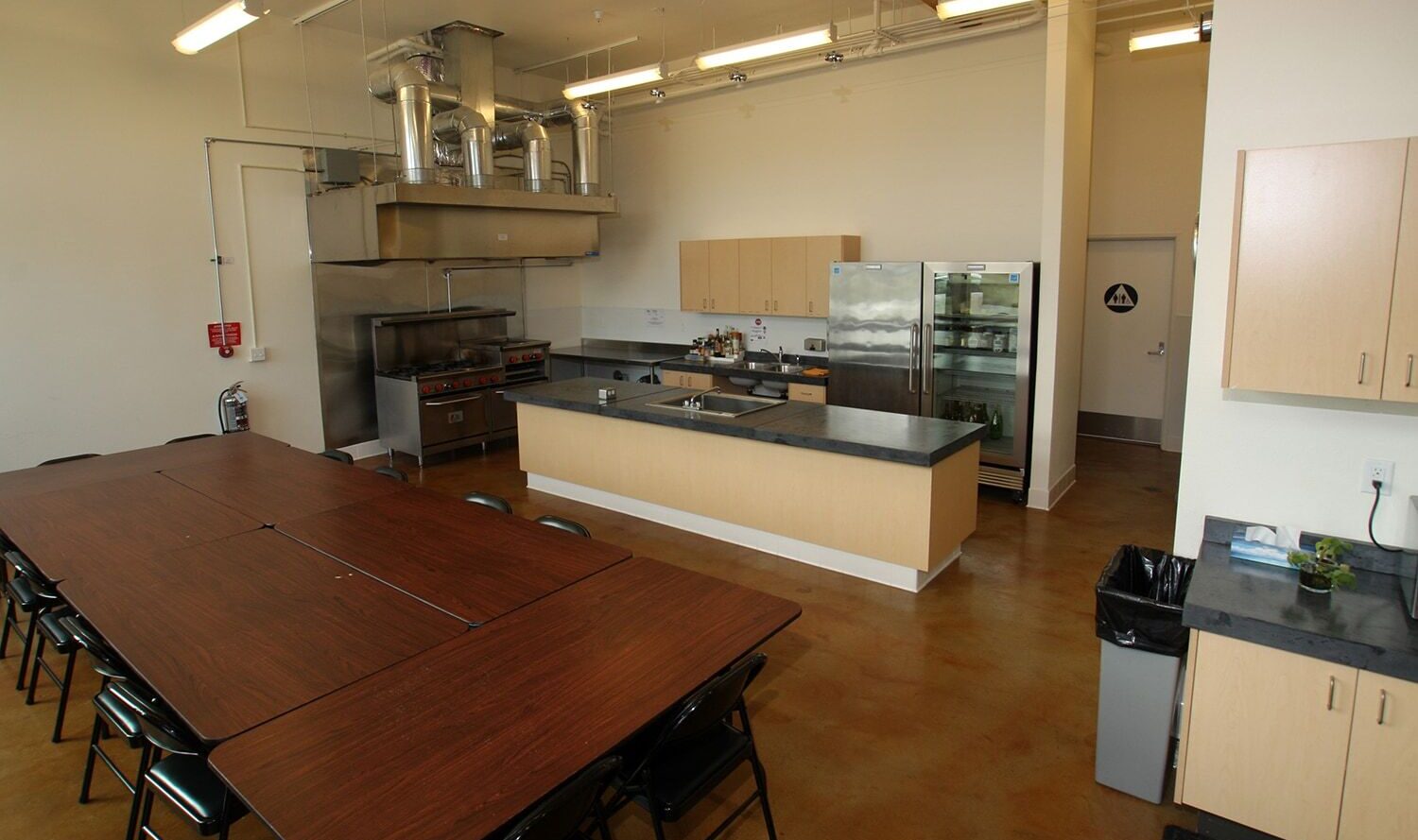 A spacious kitchen with a large central island, stainless steel appliances, and wooden cabinets. A long table with black chairs is in the foreground. Bright overhead lighting and a polished brown floor complete the room.
