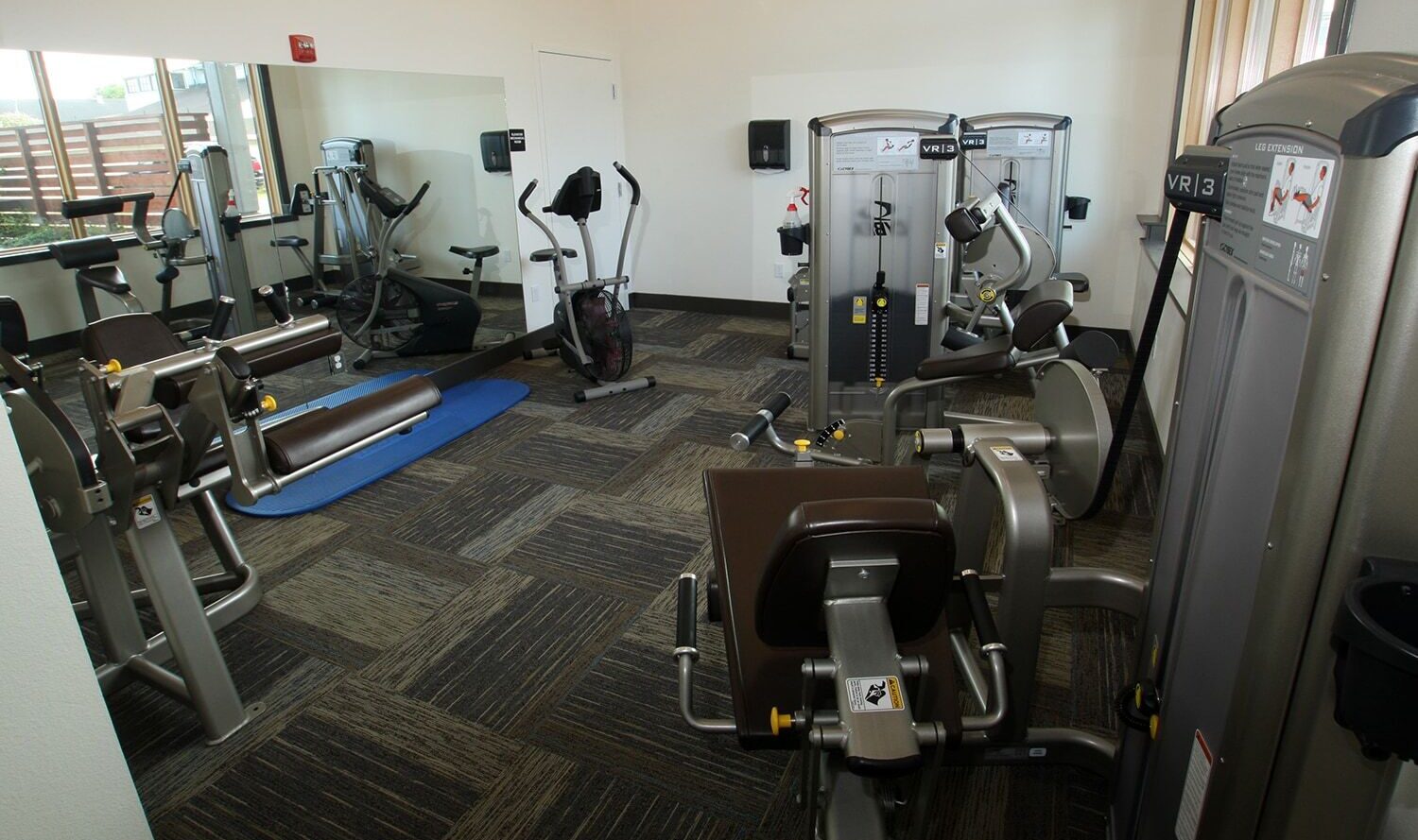 Small gym with various exercise machines, including stationary bikes, weight machines, and a blue exercise mat. The room has carpeted floors and a large mirror on one wall. Natural light enters through a window on the right.