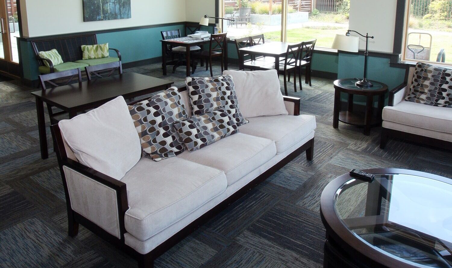 A modern lounge with a beige sofa and decorative pillows, glass-top coffee tables, and wooden chairs. Large windows provide a view of a garden outside. The room has a patterned carpet and walls adorned with minimalistic artwork.