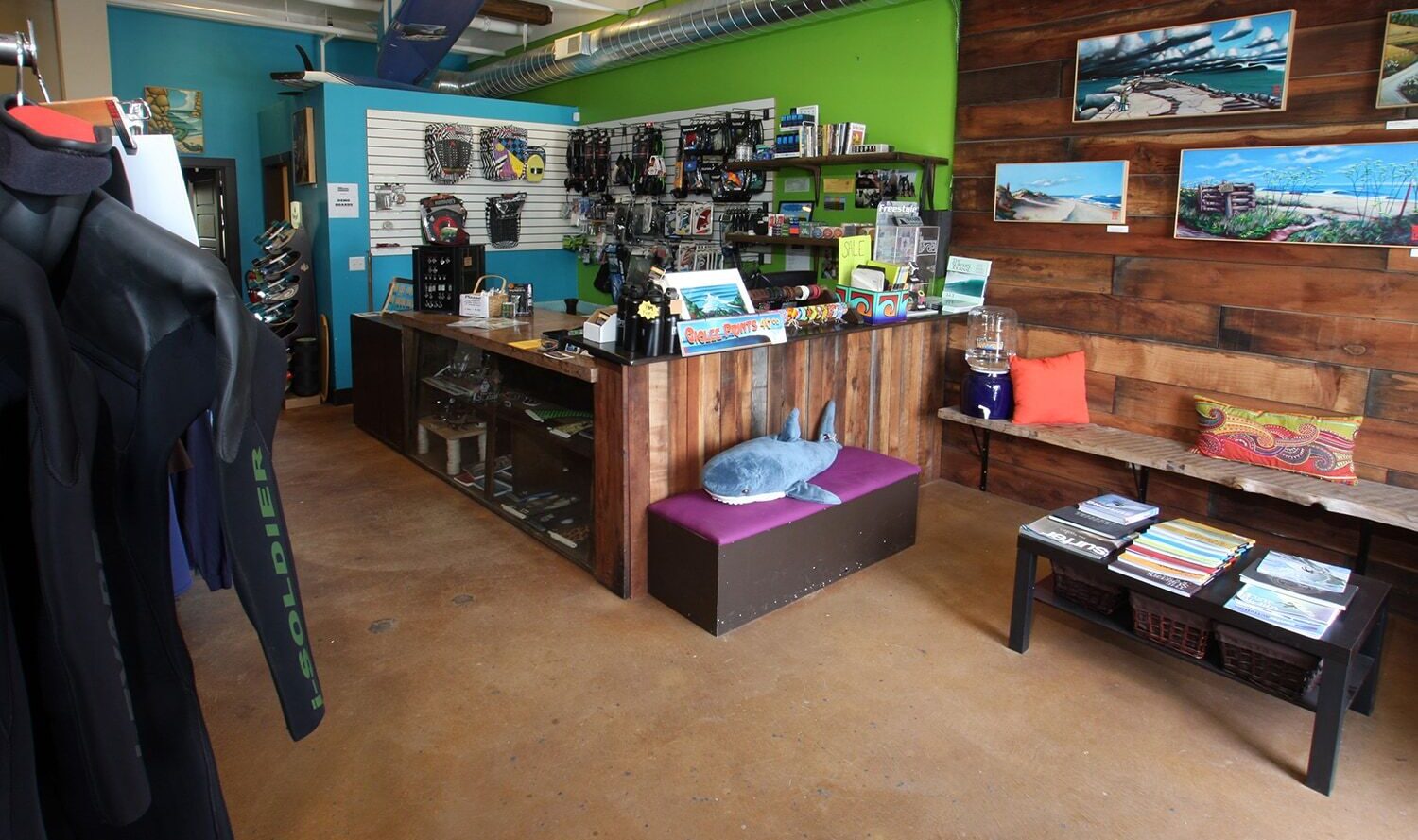 A surf shop interior with wooden decor, featuring surfboards on the wall, a counter with various gear, wetsuits hanging, and seating with colorful cushions. Bright green and blue walls enhance the vibrant atmosphere.