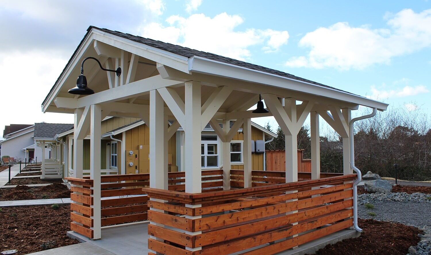 A small wooden pavilion with a dark roof and exposed beams stands outdoors. It features rustic horizontal wood paneling and a black lamp. Nearby, a yellow house and blue sky with clouds are visible.