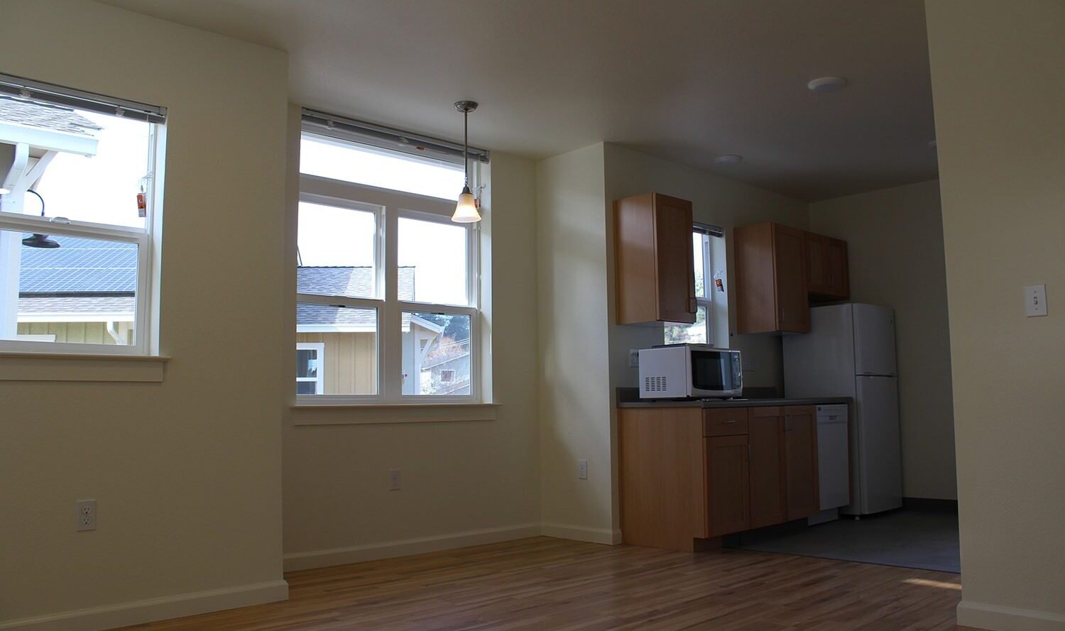 A small, empty kitchen and dining area with light wood flooring. The kitchen features wooden cabinets, a microwave, and a refrigerator. Two large windows allow natural light to illuminate the space.