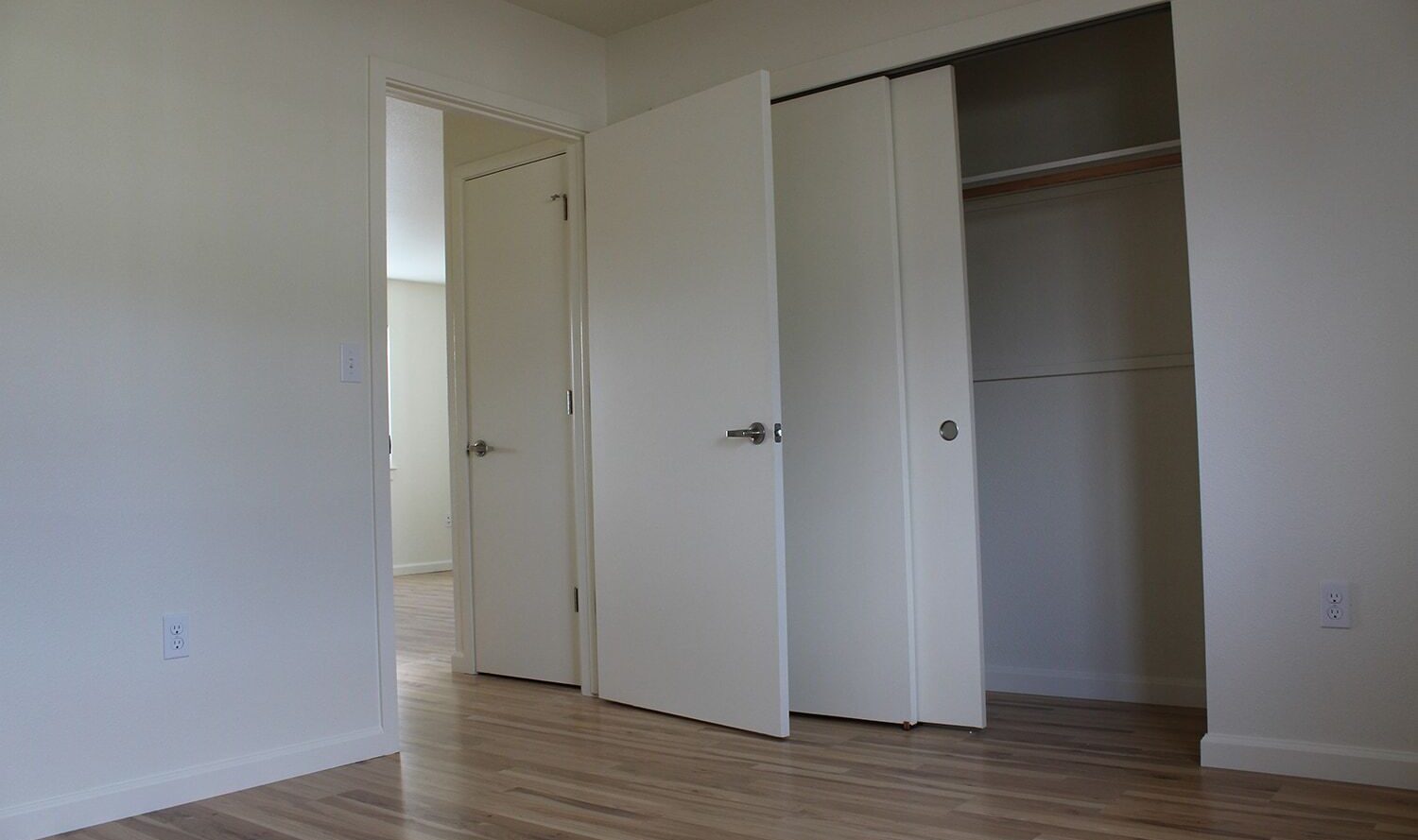 A minimally furnished room with light wood flooring, white walls, and an open closet. One door is slightly ajar, showing an adjacent room. The closet has sliding doors and a simple interior shelf.