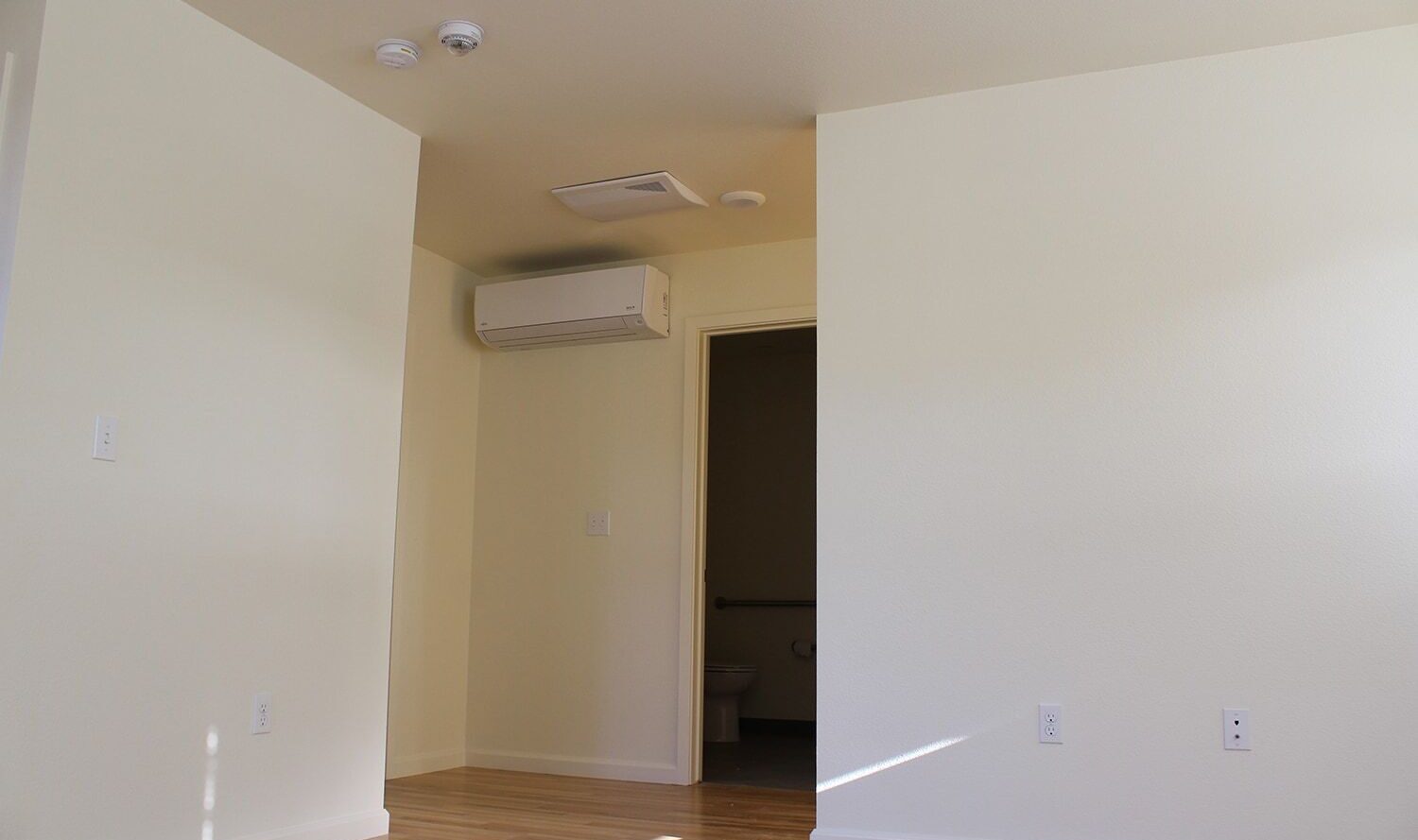 A minimalistic room with light-colored walls and wooden flooring. An air conditioning unit is mounted on the wall above a door leading to a small, partially visible bathroom.