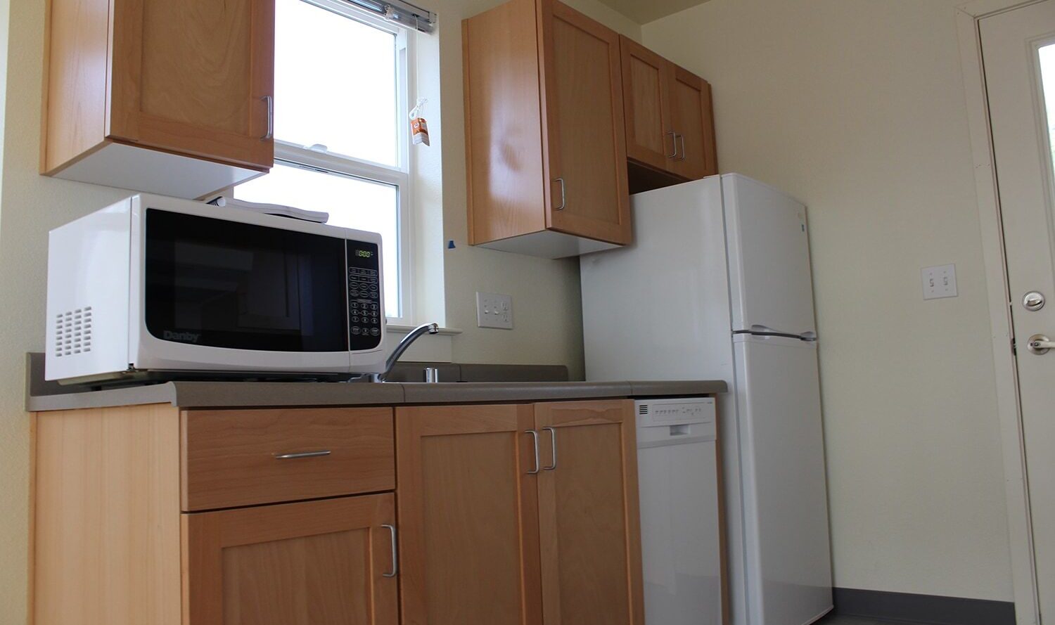 A kitchen with light wooden cabinets, a countertop with a sink, and white appliances including a microwave, dishwasher, and refrigerator. A window above the sink lets in natural light.