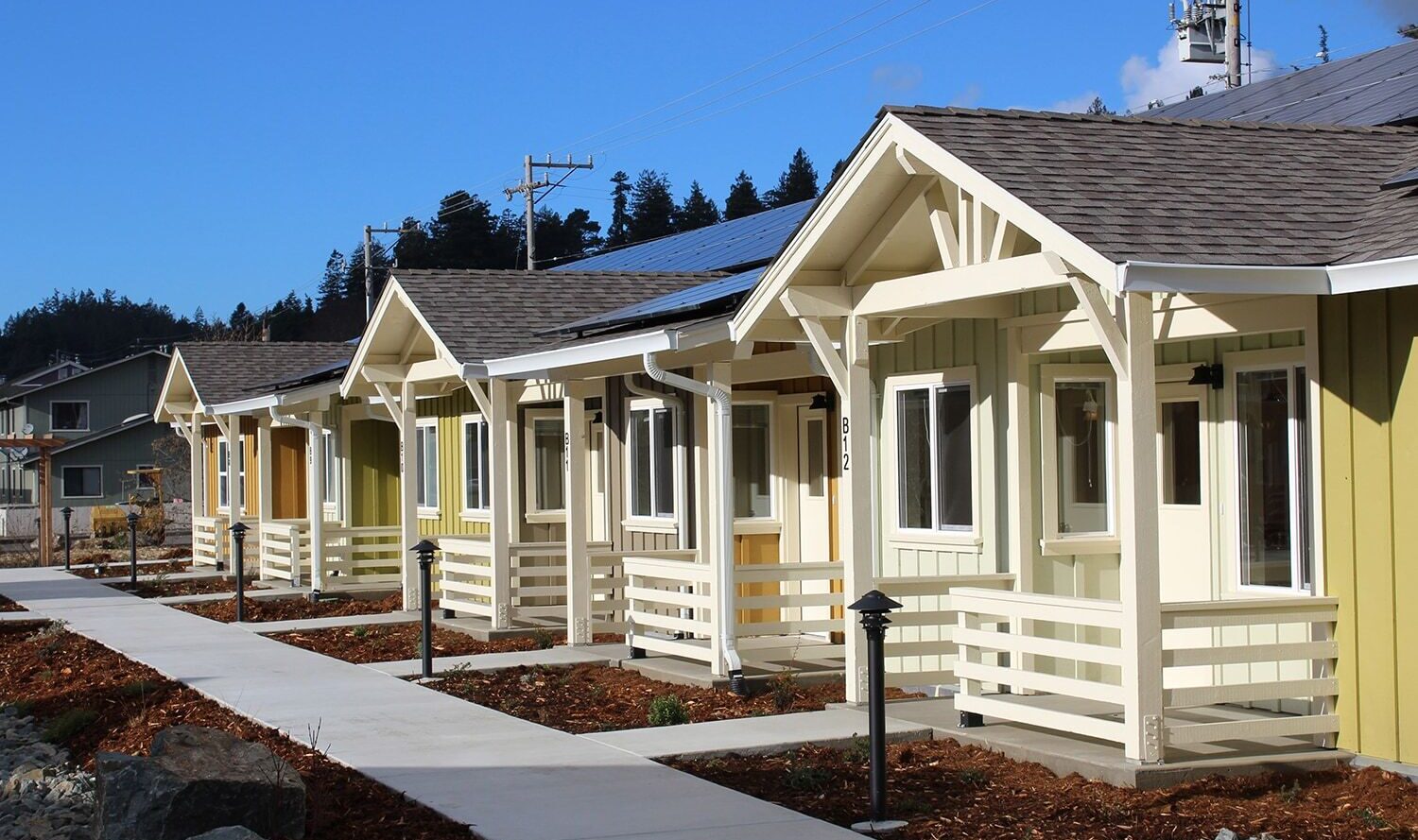 Row of small, colorful houses with white trim and solar panels on roofs, lined along a clean sidewalk. Clear blue sky and trees in the background.