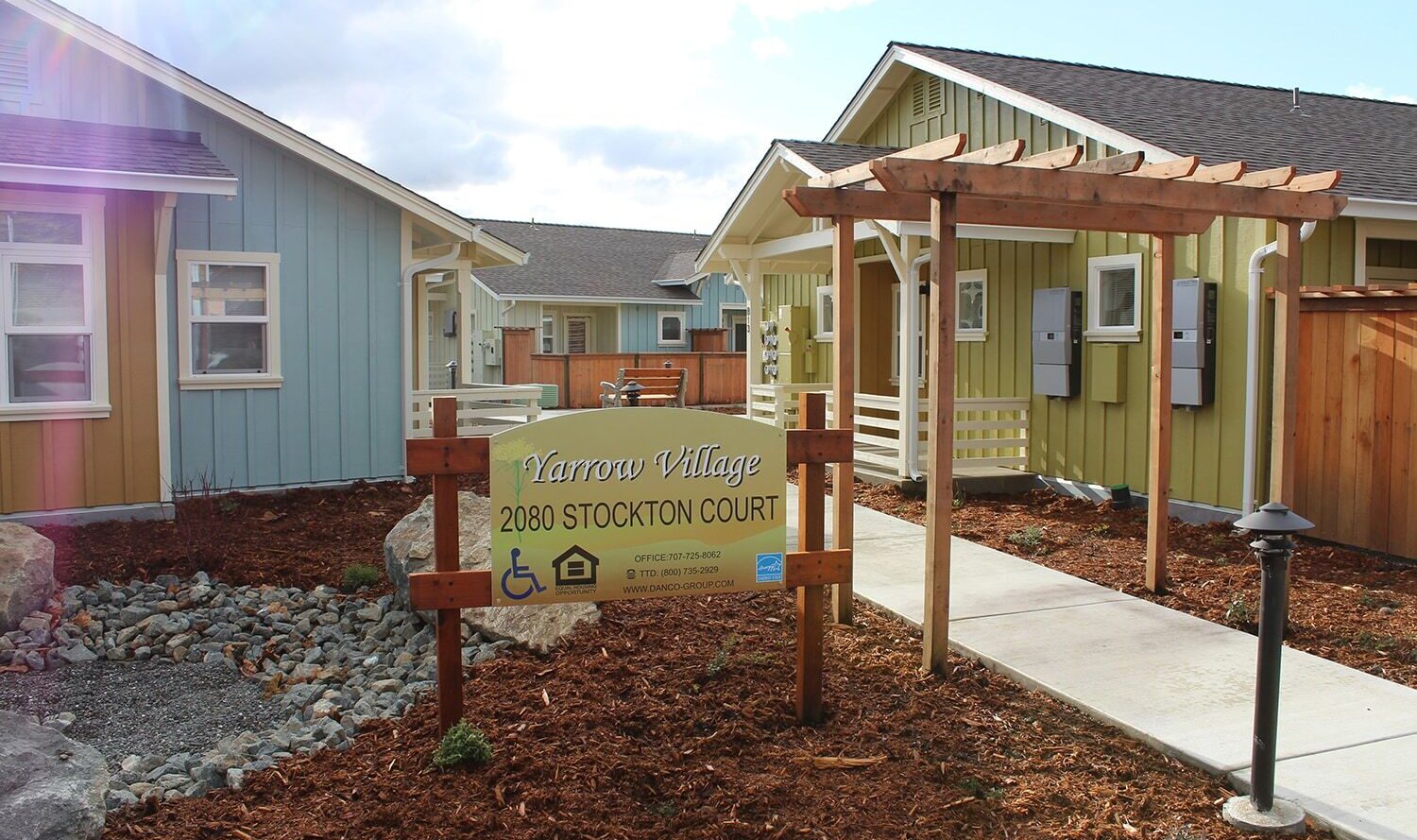 A small residential community with two single-story houses, one blue and one green. A sign reads Yarrow Village, 2080 Stockton Court with icons for accessibility and housing. A wooden archway leads to a walkway between the homes.