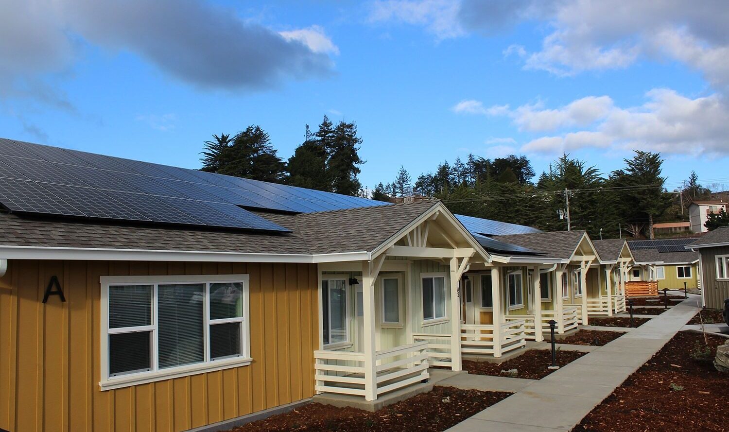 A row of modern, single-story homes with yellow and beige exteriors, each featuring solar panels on the roof. A concrete pathway runs along the front of the homes, with landscaped gardens and trees in the background under a partly cloudy sky.