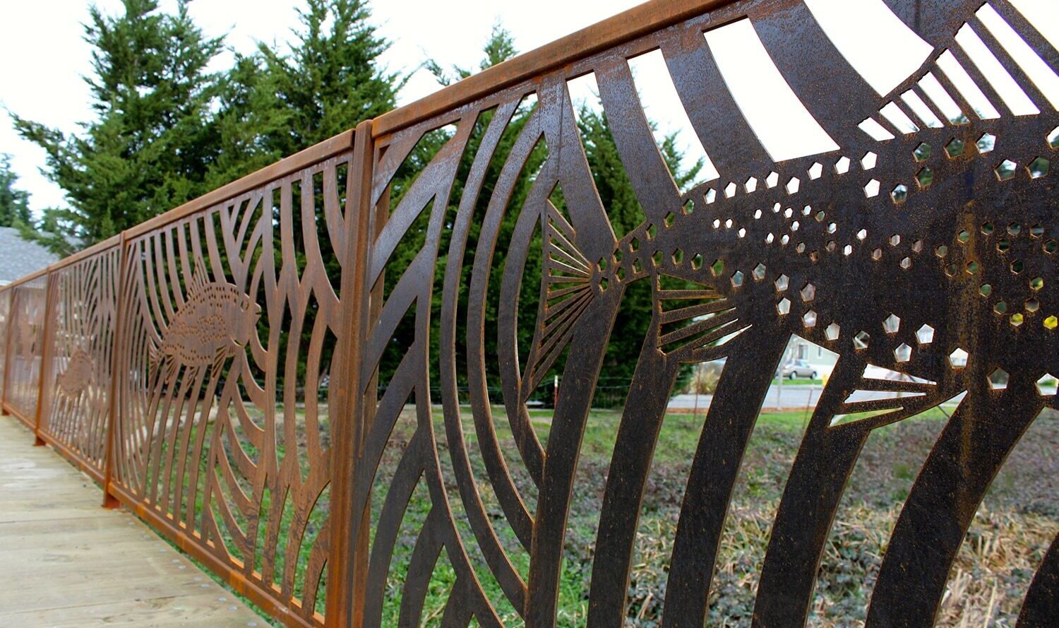 A decorative metal fence with artistic fish and crab cutouts, featuring rusted textures. The fence is situated on a wooden walkway, with evergreen trees visible in the background.