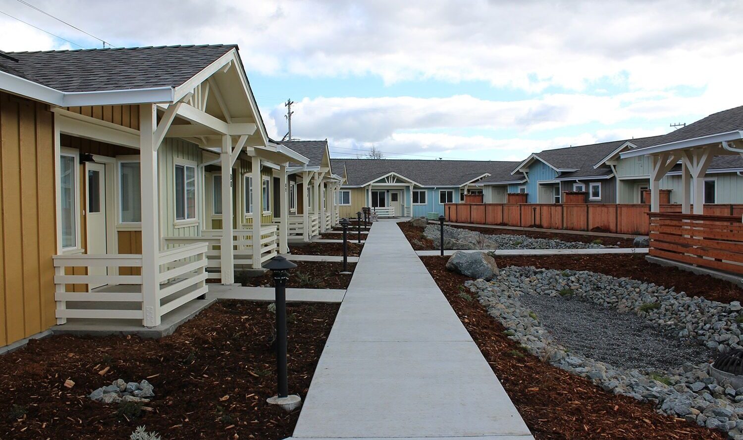 A row of modern, single-story houses with tan and blue exteriors, white trim, and covered porches. They are lined along a central concrete pathway, with landscaped gardens and wooden fences on either side under a partly cloudy sky.
