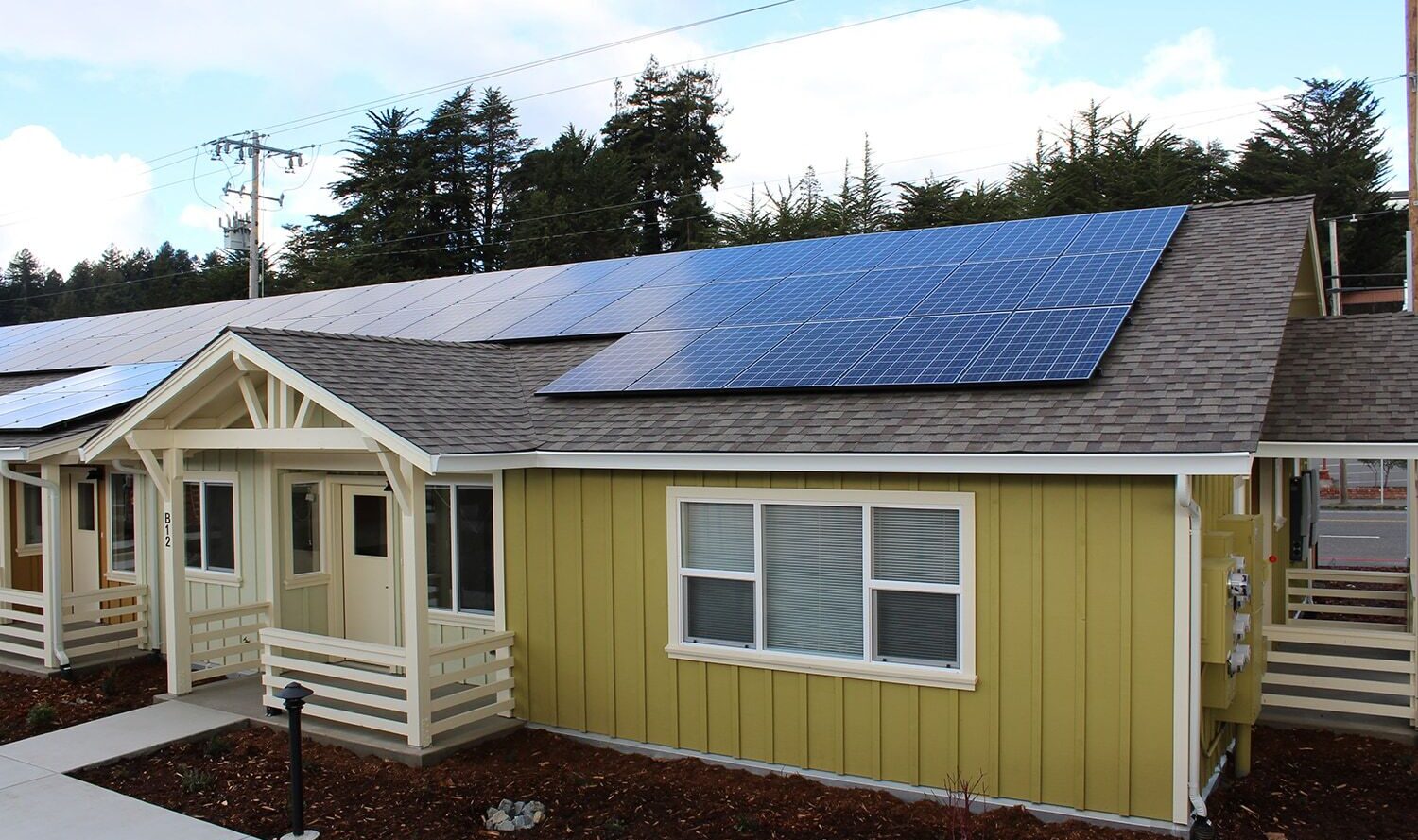 A small, yellow house with solar panels on the roof. It has white trim and a small porch. The house is part of a row of similar houses. The sky is clear with a few clouds, and there are trees in the background.