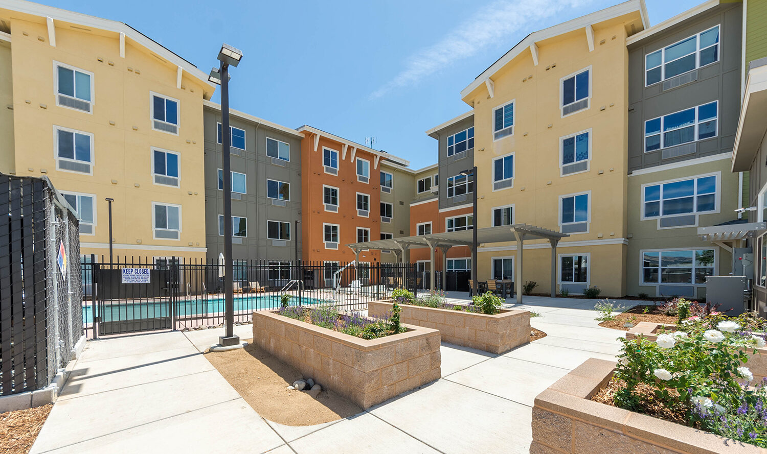 Modern apartment complex with four stories in various shades of yellow, orange, and gray. A fenced pool area is in the courtyard, surrounded by planters and pathways. The building is under a clear blue sky.