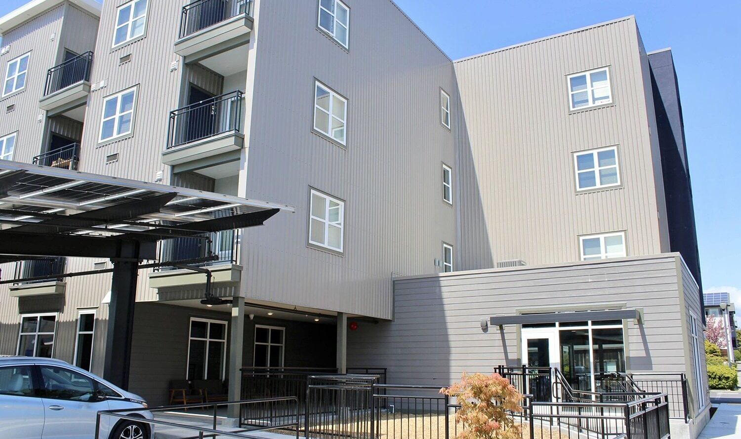 Modern four-story apartment building with balconies, featuring gray siding and numerous windows. A parking area with a silver car is partially covered by a solar panel structure. A small landscaped area with a few plants is visible in the foreground.