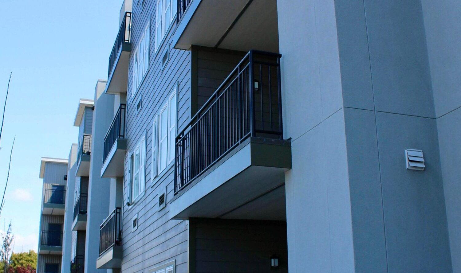 A modern apartment building with multiple stories featuring light gray and beige siding. Each unit has a balcony with black railings. The foreground shows a sign with symbols, indicating non-smoking and pet-friendly areas.