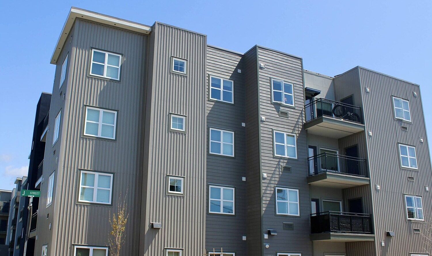 A modern, multi-story apartment building with light gray cladding and a series of balconies on one side. The sky is clear and blue, and a small tree is visible in front of the building.