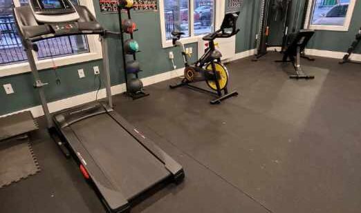 A gym room with a treadmill, stationary bike, and weight lifting equipment. Dumbbells are stacked in the corner near a rack with other exercise gear. Two large windows allow natural light inside, highlighting the exercise charts on the walls.