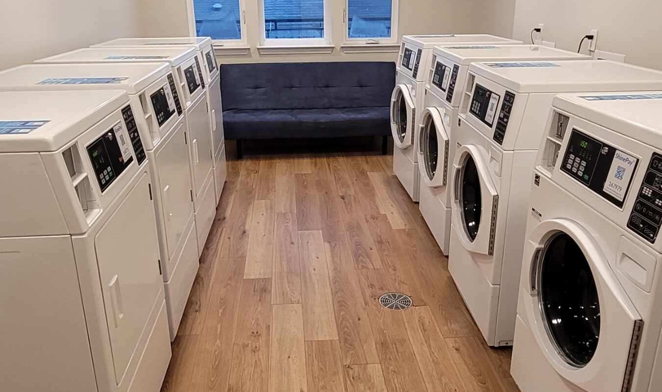 A bright laundry room with wooden floors featuring two rows of white washing machines and dryers. A small couch is placed in front of large windows at the end of the room, letting in natural light.