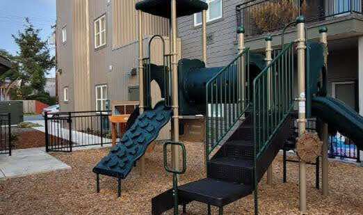 A playground with climbing equipment and slides is surrounded by wood chips, situated adjacent to a multi-story building. Theres a safety fence nearby and some trees and greenery in the background.