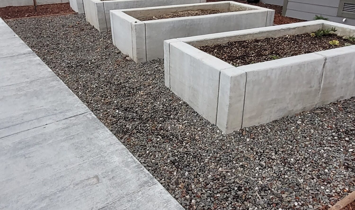 Concrete planters filled with gravel and some soil are placed beside a modern building. A sidewalk runs alongside, and a car is visible in the background. The setting appears to be a residential area.